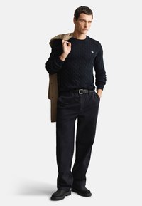 Tengerészkék kötött pulóver, fekete nadrág, fekete csizma, vállára vetett bézs kabát. Egyszerű kialakítás, texturált anyag.