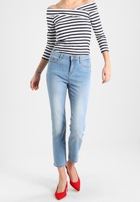 Jeans de mezclilla azul con un ajuste slim y dobladillo ligeramente recortado. Combinados con una blusa a rayas sin hombros y zapatos de tacón rojo.