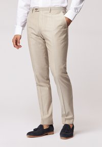 Beige Anzughose mit flacher Front, seitlichen Taschen und schmaler Passform. Getragen mit schwarzen Wildleder-Pantoletten mit Schleifchen.