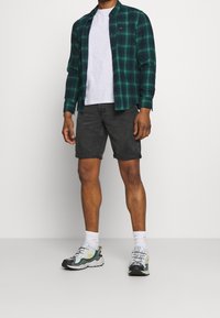 Chemise en flanelle à carreaux verts et noirs portée sur un t-shirt gris, associée à un short en jean noir et des baskets grises avec des accents jaunes et des chaussettes blanches.