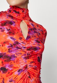 Robe florale rouge avec un col haut, une découpe en trou de serrure et un ruissellement texturé. Les couleurs comprennent des accents violets et noirs sur un fond vibrant.