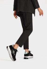 Sneaker nere realizzate con materiali in pelle e camoscio, dettagli argentati sul tallone, suola bianca spessa e lacci neri. Indossati con pantaloni neri su misura.