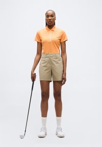 Γυναίκα με πορτοκαλί πόλο, μπεζ σορτς, λευκά παπούτσια, κρατώντας μπαστούνι του γκολφ, στέκεται μπροστά σε απλό λευκό φόντο.