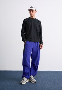Schwarzes Langarmoberteil, lila Baggyhosen und graue Mütze. Strukturierte Sneakers mit dicker Sohle. Einfaches Design und kontrastierende Farben.
