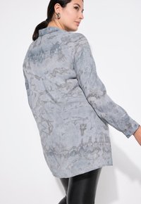 Šedá dlouhá tričko s tie-dye vzorem, s límečkem, volným střihem a mírně zakřiveným spodním lemem, vyrobené z lehkého materiálu.