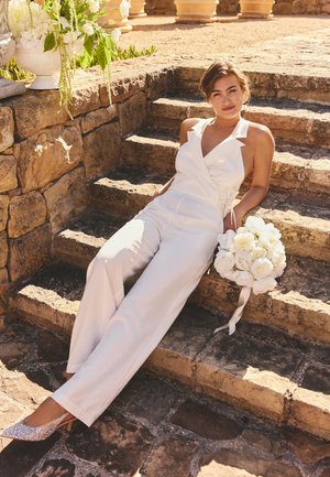 Vrouw in een wit mouwloos jumpsuit die een wit boeket bloemen vasthoudt, zittend op stenen trappen met decoratieve vazen op de achtergrond.