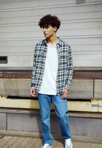 Geruite shirt in groen en wit, witte t-shirt eronder, blauwe spijkerbroek en witte sneakers. Denim textuur en knoopdetails zichtbaar.