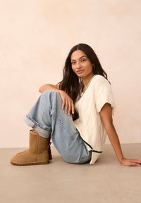 Krém színű kötött pulóver, világoskék bő szárú farmer és barna velúr bokacsizma. A modell semleges háttér előtt ül.