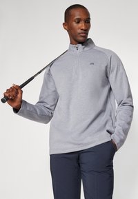 Grå, langermet genser med halv glidelås og brodert logo, sammen med mørke bukser, holdt en golfklubbe i den ene hånden.