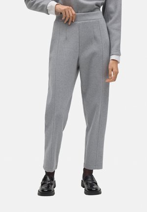 Pantalones grises ajustados con un acabado texturizado, que cuentan con pliegues en los laterales y una cintura alta. Combinados con mocasines negros y una blusa con puños blancos.