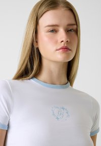 Weißes eng anliegendes T-Shirt mit hellblauem, geripptem Kragen und Ärmeln; in der Mitte mit einem filigranen blauen Blumenmonogramm versehen.