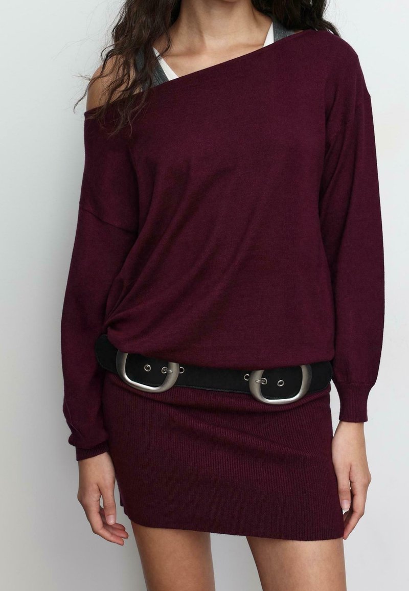 Femme portant une robe en maille bordeaux à épaules dénudées, manches longues, avec une large ceinture noire à deux boucles argentées.