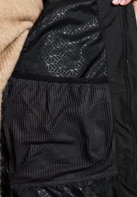 Das Innenfutter der schwarzen Jacke zeigt ein geometrisches Muster und strukturierte Mesh-Bereiche. Die Außenseite ist glatt, mit einem weichen, beigefarbenen Stoff, der sichtbar ist.