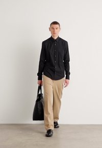 Zwarte overhemeld, beige broek en zwarte schoenen. Het model heeft een grote zwarte handtas bij zich. Glad stof en op maat gemaakte pasvorm.