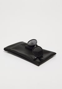 Lunettes de soleil noires avec monture ronde reposant sur une pochette en cuir noir embossée du nom de la marque "QUAY." Texture lisse, design minimaliste.
