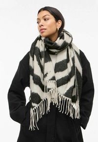 Mustavalkoinen huivi, jossa rohkea kuviointi. Reunassa tupsut ja pehmeä, kodikas rakenne, mallin kaulan ympärille kiedottuna.