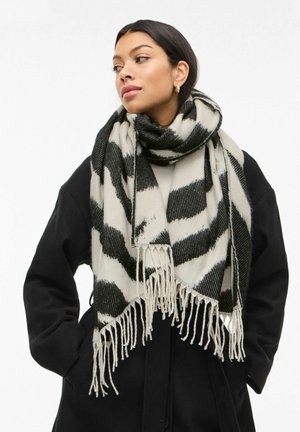 Sort og kremfarget skjerf, vevd med et dristig mønster. Har frynsete kanter og en koselig tekstur, drapert rundt halsen til en modell.