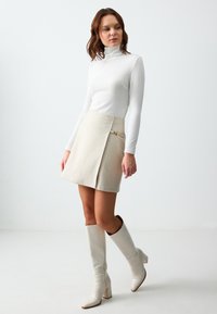 Beige rok met een A-lijn vorm en zijden gouden hardware details, gecombineerd met een aansluitende witte coltrui met lange mouwen en hoge witte laarzen.