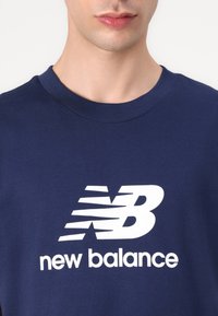 Tummansininen puuvillainen t-paita, jossa on suuri valkoinen New Balance -logo ja vinoja raitoja, pyöreä pääntie ja lyhyet hihat.