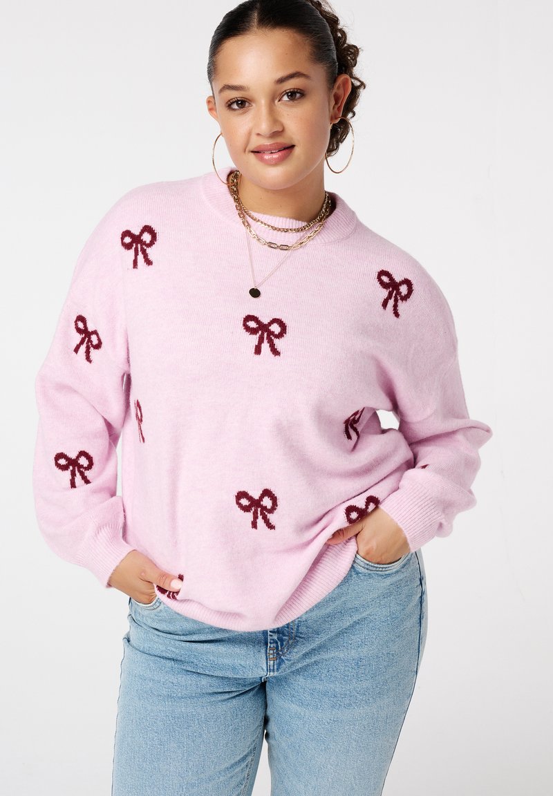 Vrouw met hoepeloren, gekleed in een roze sweater met donkerrode strikpatronen en lichtblauwe jeans, poserend met haar handen in haar zakken.