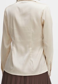 Blouse en satin beige à manches longues et col, présentant une texture lisse et une coupe ajustée, associée à une jupe plissée marron.