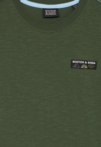 Olive green short-sleeve t-shirt with a round neckline, featuring a black embroidered logo patch with mountain and nature motifs.