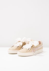 Baskets en cuir beige avec une semelle blanche texturée, ornées de grands nœuds en satin blanc pour les lacets et d'un charme décoratif sur le côté.