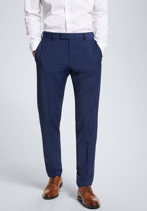 Mand iført marineblå dressbukser, hvid skjorte stoppet ind, hænder i lommerne og brune lædersko på en ensfarvet baggrund.