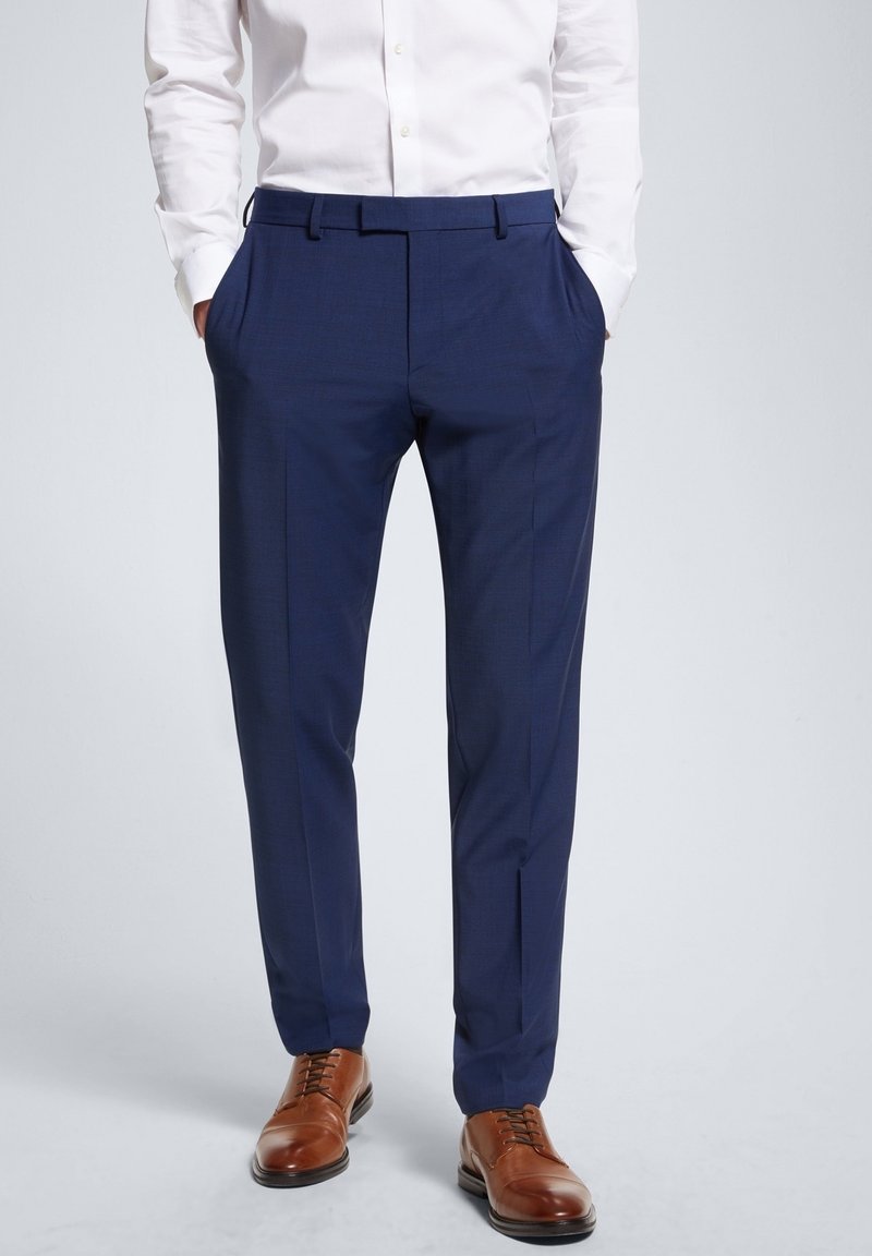 Homme portant un pantalon de costume bleu marine, une chemise blanche rentrée, les mains dans les poches, et des chaussures de ville en cuir marron sur un fond uni.