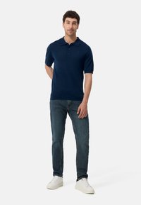 Marinblå kortärmad pikétröja med ribbad krage, tvåknapps knappslå och en subtil logotyp, kombinerad med åtsittande blåjeans och vita sneakers.