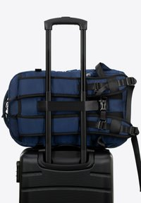 Marineblauwe duffel tas vastgemaakt aan zwarte koffer met wielen, met gewatteerde banden, ritssluiting en een textuurmateriaal met een ruitpatroon.