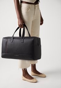 Black textured leather duffel bag with dual handles, zipper, and a red accent stripe. Worn with cream-colored, frayed hem trousers and flats.