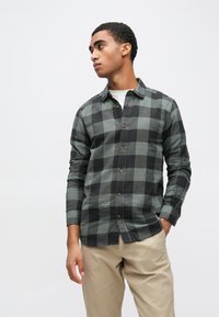 Camisa de flanela às quadrados em verde escuro e preto, com design de abotoar, mangas compridas e camiseta branca por baixo.