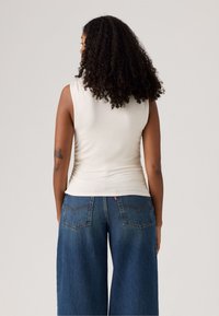 Top sin mangas de color crema con un diseño ajustado, combinado con pantalones vaqueros de pierna ancha de color azul oscuro que cuentan con bolsillos traseros y un sutil detalle de costura.
