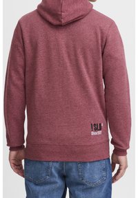 Rode hoodie met een textuurstof. Bevat een logo op de onderrug en ribgebreide manchetten. Draagt samen met blauwe spijkerbroeken.