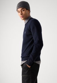 Navy knit sweater with a crew neckline, made of soft fabric. Worn over a light gray collared shirt, complemented by a gray ribbed beanie.