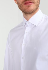 Man in een witte overhemd met een puntige kraag en een subtiele "CK"-logo op de borst.