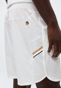 Λευκά αθλητικά σορτς με ελαστική μέση, πλαϊνή τσέπη, μαύρες και καφέ ρίγες και μικρό μαύρο και καφέ λογότυπο στον αριστερό μηρό.