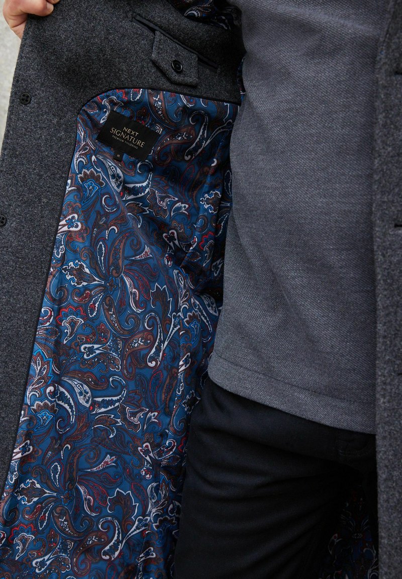 Mano sosteniendo un abrigo oscuro gris abierto, revelando un forro de pañuelo azul con un patrón paisley y una etiqueta "Next Signature" en el interior.