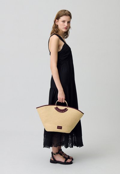 Sac en osier tressé de forme courbe, avec des accents en cuir marron et une poignée ronde, tenu par une personne portant une robe noire avec des bordures en dentelle.