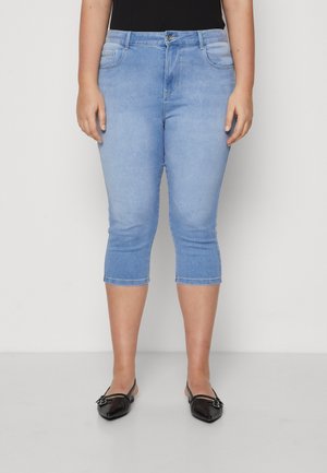 Vrouw draagt lichtblauwe cropped skinny jeans en zwarte platte schoenen met puntige neus, staand tegen een effen achtergrond.