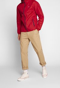 Man i röd zip-up jacka, beige upprullade chinos, vita högtoppsneakers, står med händerna i fickorna framför en enfärgad bakgrund.