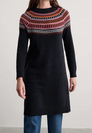 Femme portant une longue robe-pull noire en maille avec un empiècement à motif multicolore et un jean bleu sur un fond uni.