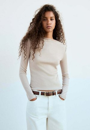 Jeune femme aux longs cheveux bouclés, portant un haut beige à manches longues, un pantalon blanc et une ceinture marron, les mains dans les poches.