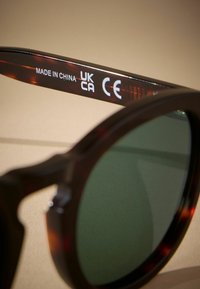 Tortoiseshell sunčane naočale sa zelenim staklima, sa zakrivljenim dizajnom i ugraviranim brendiranjem na unutrašnjem dijelu strani, uključujući oznaku "Made in China" i CE oznake.