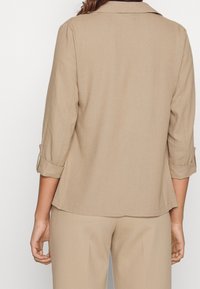 Beżowa dopasowana marynarka z klapami z wycięciami, rękawami do trzech czwartych długości podwiniętymi na mankietach oraz gładką, teksturowaną tkaniną.