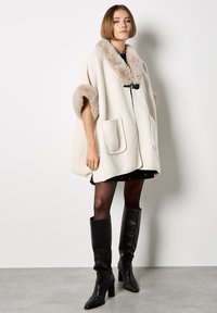 Jonge vrouw in een roomkleurige cape met bonttrim, zwarte jurk, doorschijnende panty’s en zwarte leren laarzen met hakken tot de knie, staand binnen.