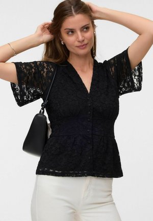 Femme portant un chemisier court en dentelle noire et un pantalon blanc, ajustant ses cheveux avec ses deux mains, portant un petit sac à épaule noir.