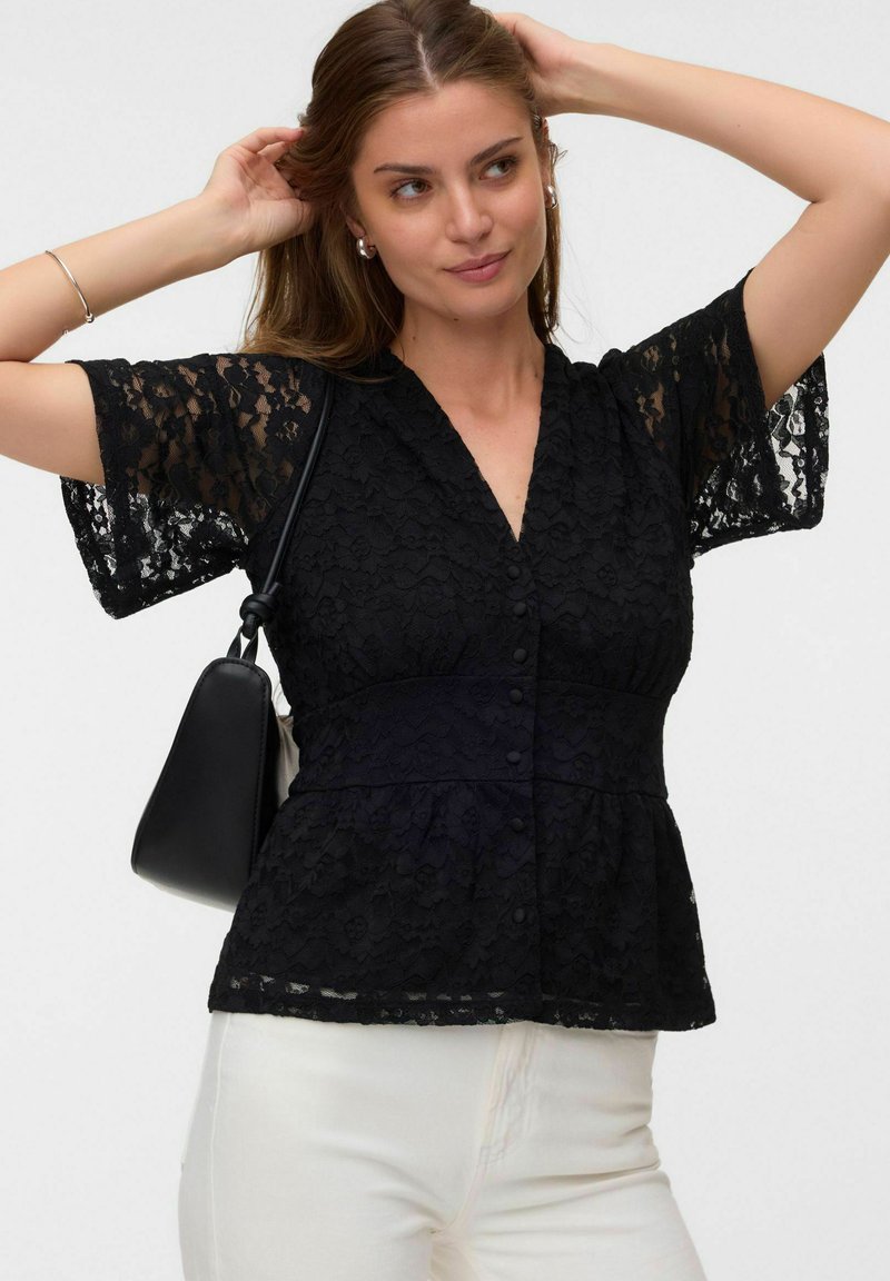 Femme portant un chemisier court en dentelle noire et un pantalon blanc, ajustant ses cheveux avec ses deux mains, portant un petit sac à épaule noir.