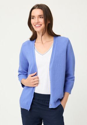 Donna sorridente, indossa un cardigan azzurro chiaro con bottoni sopra una maglietta bianca con scollo a V, pantaloni scuri, mano in tasca, su uno sfondo neutro.